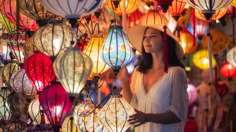 A visitor reaching up among dozens of lit silk lanterns hanging in a shop, blue, pink and red globes crowding the frame