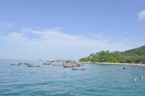 Fishing boats anchored in calm water below a steep green hillside and a scatter of village roofs