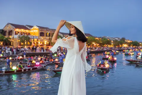 A woman in a white dress and sun hat on the Hoi An riverbank at dusk, lantern-hung boats crossing the water behind her
