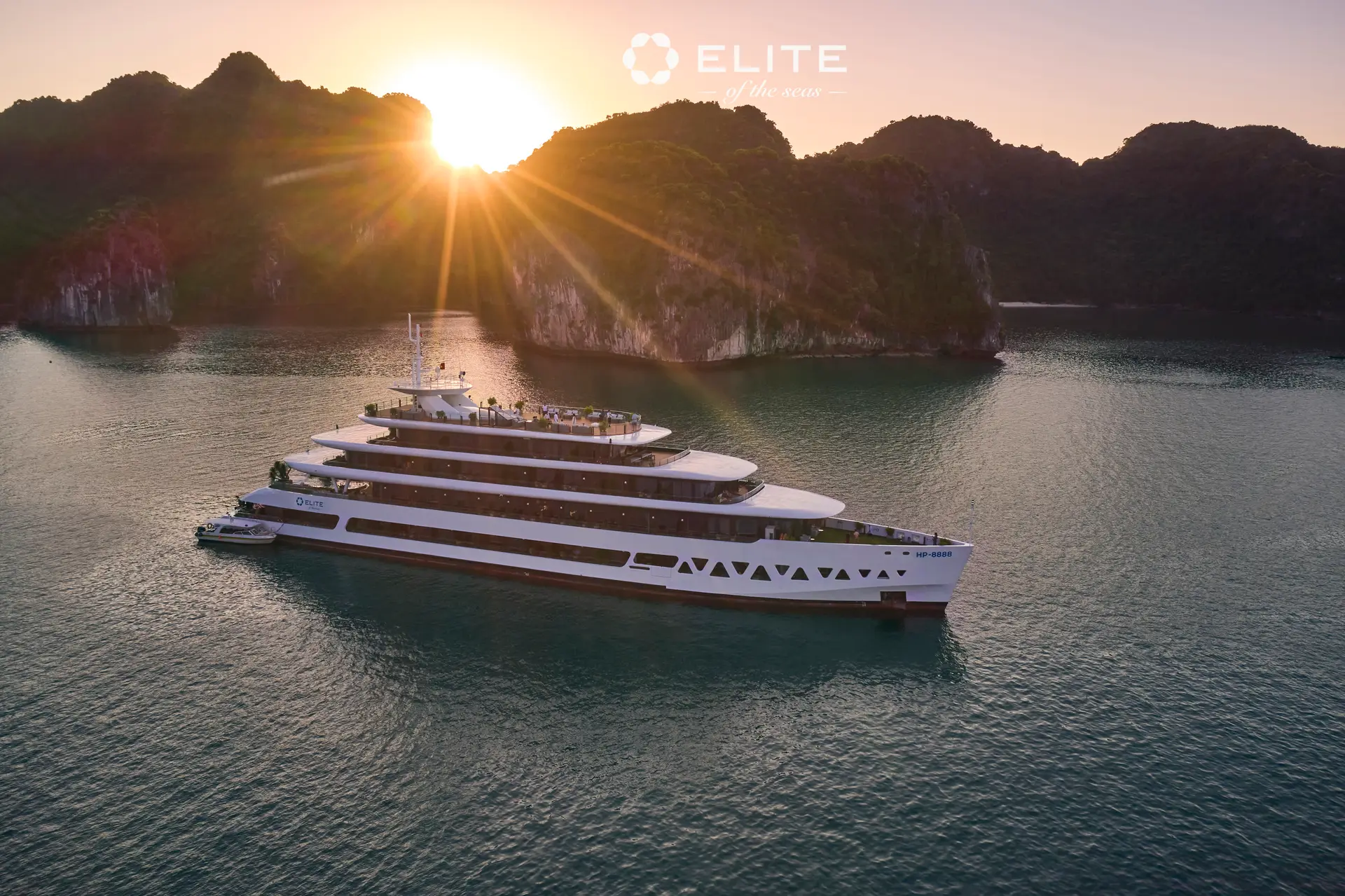 Elite of the Seas broadside on still water with the sun breaking over the karst ridge behind her