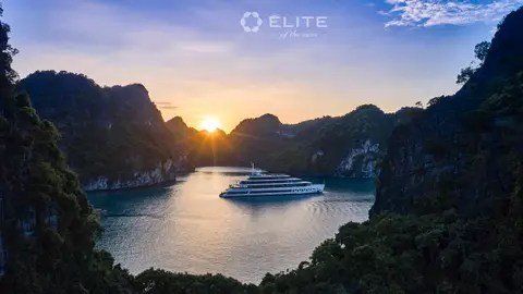 The ship at anchor between two karsts as the sun sets down the channel behind her
