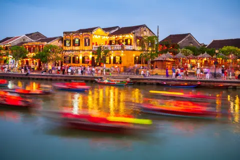 The Hoi An waterfront at dusk, lit shophouses along the bank and boats drawing coloured trails across the river