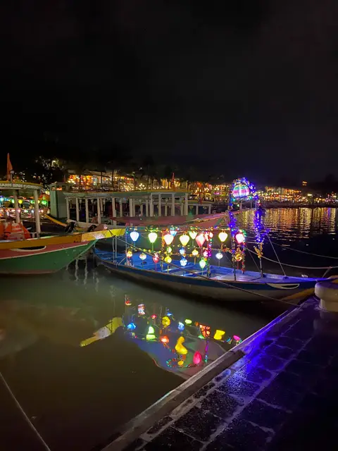 Row of moored boats lit with red and blue lanterns along a riverbank after dark