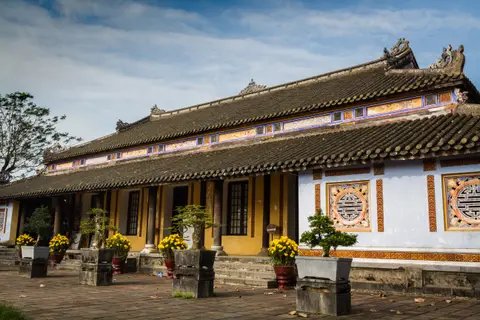 Ochre and red palace hall with a tiled roof and carved timber doors, potted plants along the terrace