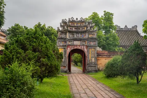 Ornate arched gateway with mosaic-tiled roofs, seen across a lawn and clipped shrubs