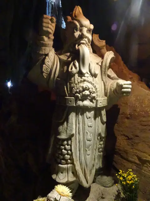 Carved stone guardian figure set against the dark rock wall of a cave, lit from one side