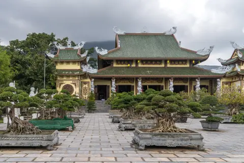 Tiered pagoda hall with sweeping tiled roofs, approached across a paved court lined with bonsai