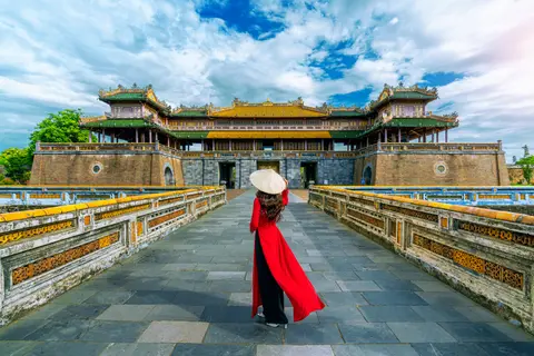 Une femme en ao dai rouge marchant sur l'avant-cour carrelée de la Cité impériale de Hue vers une porte à toit jaune