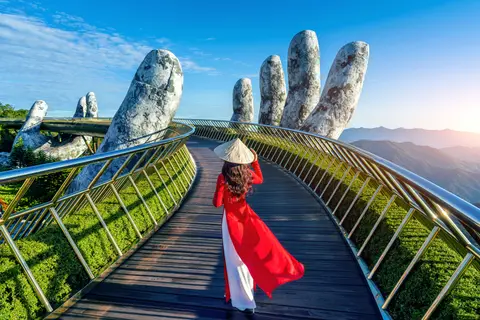 Una mujer con ao dai rojo caminando por el Puente Dorado, el pasarela dorada curvando entre crestas boscosas