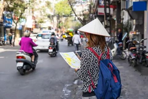一位戴着斗笠的旅行者在河内繁忙的街道上阅读纸质地图，身后是拥挤的摩托车和店铺门面